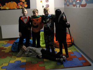 Pięcioro uczniów klasy czwartej pozuje do wspólnego zdjęcia. Czworo chłopców stoi obejmując się ramieniem. Uczeń stojący po prawej stronie ma zasłoniętą twarz a w dłoni trzyma replikę kosy. Przed nimi na podłodze leży oparty na jednym ramieniu czwarty uczeń.