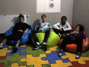Czterech uczniów rozsiada się na kolorowych pufach ułożonych na dywanie. Jeden z nich trzyma przed sobą kufel z żółtym płynem. Obok niego siedzi uczeń z chustą na twarzy trzymający w dłoni replikę pistoletu. Dalej siedzi uczeń bez stroju a na końcu chłopiec w czarnym stroju z przyłbicą na głowie.