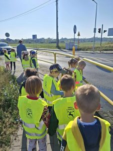 Uczniowie oddziału przedszkolnego stoją obok siebie w odblaskowych kamizelkach na chodniku przy skrzyżowaniu. Dzieci oddzielają od ulicy żółte barierki.