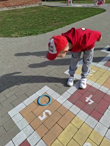 Uczeń oddziału przedszkolnego nisko pochyla się patrząc na niebieski krążek, leżący na klasach namalowanych na szkolnym boisku. Krążek leży na polu o nr 5 a chłopiec ubrany jest w czerwoną bluzę a na głowie ma czerwoną czapkę z daszkiem.