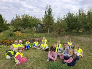 Uczniowie oddziału przedszkolnego siedzą w grupie na trawie przed sadem owocowym. Dzieci mają na sobie odblaskowe kamizelki.