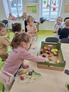 Uczennica oddziału przedszkolnego sięga ręką do tekturowej skrzynki, w której leżą jabłka i gruszki. W tle widać chodzących po sali jej kolegów i koleżanki.