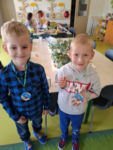 Dwóch uczniów oddziału przedszkolnego pozuje do wspólnego zdjęcia. Obaj mają zawieszone na szyi medale. Jeden z nich podnosi swój medal w dłoni. Chłopcy się uśmiechają.