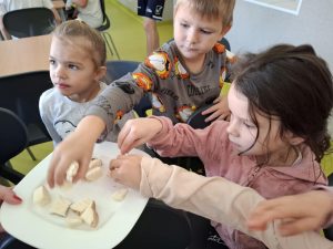 Na zdjęciu widocznych jest troje uczniów z oddziału przedszkolnego. Dwójka z nich sięga na trzymanego w powietrzu talerza po kawałki buraka cukrowego. Na pierwszym planie widoczna jest również ręka innego dziecka wyciągnięta nad talerzem.