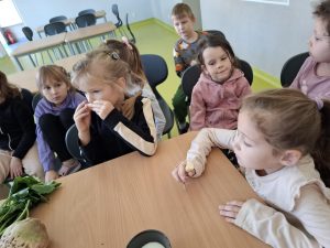 Uczniowie oddziału przedszkolnego siedzą przy szkole. Jedna z dziewczynek trzyma przy nosie w obu dłoniach kawałek buraka cukrowego.