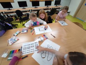 Na pierwszym planie widoczne są dwie uczennice siedzące przy dużym stole. Przed nimi leży kartka papieru nad którą jedna z uczennic trzyma dłoń. Po kartce porusza się kula znajdująca się pod dłonią dziewczynki. Koleżanka uważnie przygląda się sytuacji. Obok siedzi dziewczynka spoglądająca w ich stronę. Na stole leżą dodatkowo kolorowe pisaki oraz kartki papieru z kodami i liniami.