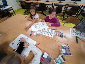 Na pierwszym planie widoczne są dwie uczennice siedzące przy stole. Przed dziewczynkami leżą kartki papieru z kodami oraz z rysunkiem zamkniętej pętli. jedna z dziewczynek patrzy w bok a druga przed siebie. Na stole leżą dodatkowo opakowania z pisakami. Na przeciwko uczennic częściowo widoczna jest dwójka uczniów rysująca na kartce papieru schemat.