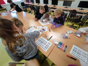 Uczniowie siedzą przy długim stole. Przed nimi leżą kartki papieru z kodami oraz kolorowe pisaki. Część z dzieci pochyla się nad kartkami i rysuje na nich wzory opisane kodami.
