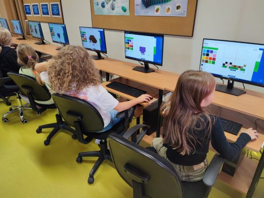Cztery uczennice siedzą na krzesłach obrotowych przy stanowiskach komputerowych. Na monitorach ustawionych przed dziewczynkami widoczne są obrazki utworzone z kolorowych kwadracików. Każda z dziewczynek trzyma dłoń na myszce ustawionej obok klawiatury.