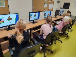 Cztery uczennice siedzą na krzesłach obrotowych przy stanowiskach komputerowych. Na monitorach ustawionych przed dziewczynkami widoczne są obrazki utworzone z kolorowych kwadracików. Każda z dziewczynek trzyma dłoń na myszce ustawionej obok klawiatury.