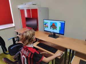 Uczeń siedzi na krześle obrotowym przy stanowiskach komputerowych. Na monitorze ustawionym przed nim widoczne są obrazki utworzone z kolorowych kwadracików. Chłopiec trzyma dłoń na myszce ustawionej obok klawiatury.
