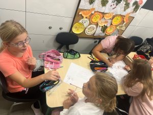 Cztery uczennice siedzą przy wspólnym stole i pracują nad własnymi plakatami. Przed nimi leżą kartki papieru oraz piórniki z przyborami do rysowania. Dwie dziewczynki pochylają się z pisakami w dłoniach nad jedną z kartek. Podczas gdy dwie pozostałe patrzą w swoim kierunku.