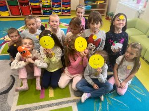 Uczniowie świetlicy siedzą obok siebie na dywanie. Część z dzieci zasłania sobie twarze żółtymi papierowymi kołami z narysowanymi uśmiechniętymi buźkami. Inne dzieci mają przyklejone do bluzek mniejsze kółka z uśmiechami. Dzieci pozują razem do zdjęcia.