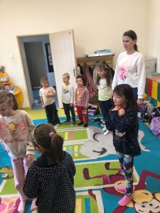 Uczniowie ze szkolnej świetlicy stoją wraz z Panią wychowawczynią na dywanie w luźnych odstępach. Część z nich ma dłonie ułożone wzdłuż ciała. Jedna z dziewczynek przytula pod szyją czerwonego pluszaka.