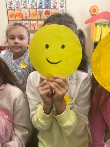 Na pierwszym planie widoczna jest uczennica, która zakrywa twarz dużym żółtym kołem wyciętym z papieru. Na kole narysowana jest uśmiechnięta buzia. Obok i w tle widać inne dzieci.