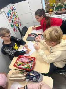 Troje uczniów pracuje przy wspólnym stole nad swoimi plakatami. Przed nimi leżą kartki papieru oraz przybory do rysowania. Dzieci pochylają się z pisakami w dłoniach nad kartkami.
