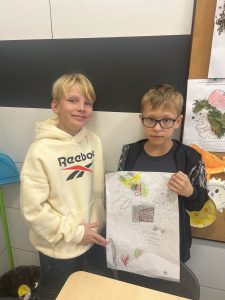 Dwóch uczniów przebywających na świetlicy pozuje do wspólnego zdjęcia. Jeden z nich trzyma przed sobą plakat wykonany z okazji Światowego Dnia Zdrowia Psychicznego. Drugi chłopiec wskazuje dłonią w jego stronę.