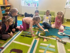 Uczniowie świetlicy siedzą na dywanie tworząc linie w kształcie litery L. Dziewczynka będąca po środku pochyla się do przodu trzymając w wyciągniętej nad dywanem ręce pluszową gęś. Na dywanie leże zamknięta przezroczysta butelka.