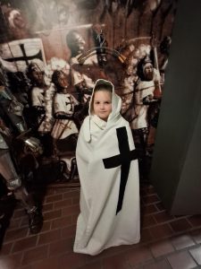 Uczeń ubrany w biały płaszcz z kapturem i dużym czarnym krzyżem stoi na tle zdjęcia uwieczniającego spotkanie krzyżaków.