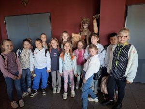 Uczniowie pozują do wspólnego zdjęcia stojąc na tle bordowej ściany, w rogu której ustawiona jest drewniana kukiełka z długim nosem trzymająca w dłoni pracę plastyczną.