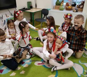 Uczniowie oddziału przedszkolnego siedzą na podłodze . Dziewczynki są ubrane w stroje krakowianek a chłopcy maja doklejone czarne wąsy. jeden z uczniów trzyma w dłoniach drewniany patyk na którym umieszczone jest popiersia kucyków. Dwie dziewczynki widoczne na pierwszym planie obejmują się.