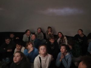 Uczniowie siedzą na podłodze i podnoszą w górę głowy. Za nimi widać ścianę kopuły na której widoczna jest mała mgiełka nad ich głowami.