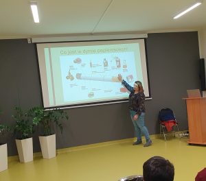 Kobieta prowadząca prelekcję stoi przy wyświetlanym na rzutniku slajdzie z grafiką dużego papierosa. Pani wskazuje dłonią na niego. Wokół widoczne są grafiki różnych substancji znajdujących się w wyrobach tytoniowych.