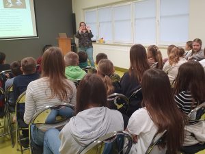 Kobieta wygłaszająca prelekcję, stoi przed uczniami trzymając w wyciągniętych na wysokość twarzy palcach wyroby tytoniowe. Przed nią widać głowy siedzących na krzesełkach uczniów.