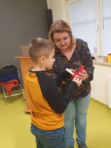 Uczeń stoi przy kobiecie prowadzącej prelekcje. Wspólnie trzymają w dłoniach czerwone urządzenie z ustnikiem. Pani tłumaczy chłopcu działanie urządzenia wskazując palcem jeden z przycisków.