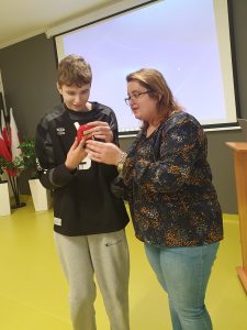 Uczeń stoi przy kobiecie prowadzącej prelekcje. Wspólnie trzymają w dłoniach czerwone urządzenie z ustnikiem. Chłopak przygląda mu się z uśmiechem na ustach.