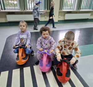 Troje uczniów oddziału przedszkolnego siedzi na pojazdach wyposażonych w kółka. Dzieci ustawione są obok siebie kaskadowo i przejeżdżają po namalowanych na podłodze toruńskiego oddziału WORD pasach.