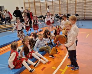 Uczniowie oddziału przedszkolnego przygotowują się do apelu z okazji odzyskania przez Polskę niepodległości na sali gimnastycznej. Część dzieci siedzi na niebieskich materacach. Na pierwszym planie widać trzech stojących chłopców w białych koszulach z przyczepionymi czarnymi wąsami. Jeden z nich patrzy w stronę obiektywu i trzyma między nogami patyk z umieszona na końcu głową pluszowego konia. W tle widać trzy dziewczynki biegnące w stronę materacy oraz innych uczniów szkoły stojących na sali.