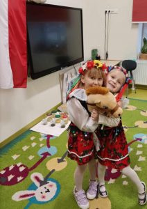 Dwie uczennice poddziału przedszkolnego stoją na zielonym dywanie i się przytulają. Dziewczynki trzymają między sobą głowę pluszowego kucyka umieszczona na drewnianym patyku.