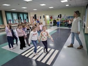 Uczniowie oddziału przedszkolnego przechodzą parami przez narysowane na podłodze sali WORDU pasy. Dzieci trzymają się w parach za dłonie. Przed nimi stoi kobieta instruująca ich jak poprawnie przechodzi się przez jezdnię.