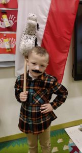 Uczeń oddziału przedszkolnego stoi trzymając jedna rękę oparta o biodro, trzymając w drugiej popiersie kucyka umieszczone na drewnianym kiju. Chłopiec ma przyklejone czarne wąsy, a za nim wisi flaga Polski.