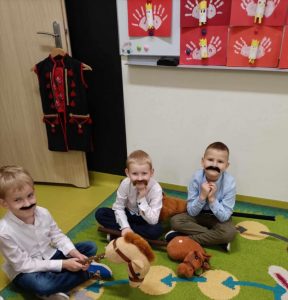 Trzech uczniów oddziału przedszkolnego siedzi na dywanie po turecku. Chłopcy mają przyklejone czarne wąsy. Jeden z nich trzyma przed sobą patyk z umieszczona na nim głową konia. Pozostali chłopcy opierają swoje głowy o przedramienia.