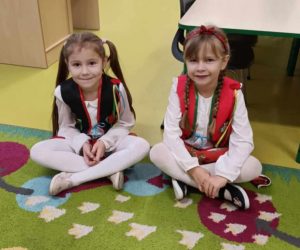 Dwie uczennice oddziału przedszkolnego siedzą na dywanie po turecku. Dziewczynki ubrane są w stroje krakowianek i uśmiechają się patrząc wprost w stronę obiektywu.