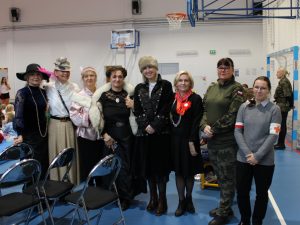 Nauczycielki wraz z Panią dyrektor pozują do wspólnego zdjęcia. Panie w większości przebrane są w stroje dam z różnych epok. Dodatkowo jedna z nich ma jest w mundurze wojskowym a druga ma założone na ramionach szarego swetra opaski krzyża medycznego i polskiej flagi.