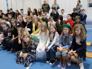 Uczniowie szkoły siedzą na ławkach na sali gimnastycznej podczas apelu. Na pierwszym planie widoczne są dziewczynki spoglądające w stronę obiektywu. Jedna z nich siedzi na podłodze.