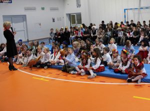 Pani dyrektor stoi na sali gimnastycznej przemawiając przez mikrofon, do siedzących naprzeciw uczniów. Część dzieci siedzi na materacach, inne na ławkach. Dzieci patrzą w stronę Pani dyrektor.