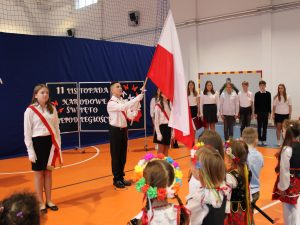 Zdjęcie pocztu flagowego stojącego przed uczniami podczas odśpiewywania hymnu. Uczeń stojący pomiędzy dwoma swoimi koleżankami trzyma przed sobą podniesioną w górę flagę Polski. Cała trójka ubrana jest na biało czarno i ma przepasane przez ramię biało czerwone wstęgi.