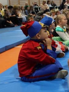 Zdjęcie siedzącego bokiem uczniami w czerwono- niebieskim stroju żołnierza. Chłopiec siedzi na materacu po turecku i opiera łokcie na kolanach. Palcami trzyma kołnierzyk swojego stroju oraz opiera na nich głowy. Patrzy przed siebie. W tle widać lekko rozmazanych innych uczniów.