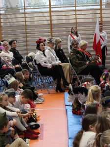 Zdjęcie siedzących na krzesłach nauczycielek obserwujących apel. Przed nimi widoczne są dwa rzędy w których siedzą uczniowie. Zdjęcie wykonane z boku.