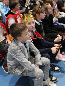 Zdjęcie uczniów siedzących na ławkach na apelu. Na pierwszym planie widoczny jest chłopiec w złotej koronie patrzący w stronę obiektywu. Pozostałe dzieci patrzą przed siebie.