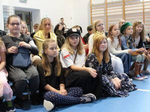 Zdjęcie uczniów siedzących na ławkach podczas apelu. Na pierwszym planie widoczne są trzy uczennice siedzące na podłodze przed ławkami. Dwie z nich patrzą w stronę obiektywu.