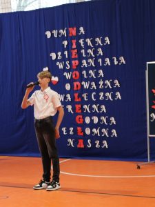 Uczeń ubrany w białą koszule z przyczepionym kotylionem i czarne spodnie stoi sam trzymając w dłoni umieszczony przed twarzą mikrofon. Na granatowym tle za nim umieszczone są pod sobą białe słowa z jedną czerwoną literą tworzące jak w krzyżówce hasło czytane pionowe. Słowa to kolejno: DUMA, PIĘKNA, DZIELNA, WSPANIAŁA, WOLNA, ODWAŻNA, WALECZNA, SUWERENNA, GODNA, NIEZŁOMNA, NASZA", a czerwono litery w tych słowach tworzą hasło "NIEPODLEGŁA".