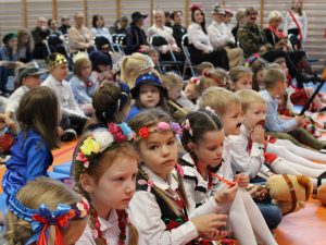Zdjęcie widowni podczas apelu. Na pierwszym planie widoczne są uczennice w strojach krakowianek siedzące obok siebie. Jedna z nich patrzy w stronę obiektywu. Tło zdjęcia jest lekko rozmazane i widać tam innych uczniów oraz nauczycieli siedzących na krzesłach.