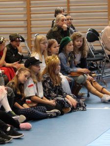 Zdjęcie uczniów siedzących na widowni podczas apelu. Na pierwszym planie widoczne są trzy dziewczynki siedzące na podłodze. Jedna z nich opiera swoją głowę o ramię koleżanki. Dzieci patrzą przed sobą. Za nimi widoczne są dwie nauczycielki siedzące na krzesłach.