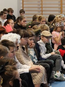Uczniowie szkoły siedzą na ławkach podczas apelu. Jeden z chłopców w jasnobrązowym kaszkiecie na głowie patrzy w stronę obiektywu. Pozostałe dzieci patrzą przed siebie.
