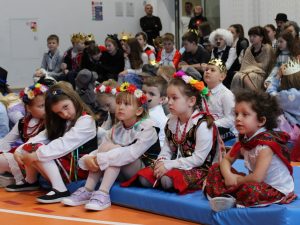 Uczniowie siedzą na materacach podczas apelu. Na pierwszym planie widoczne są dziewczynki z oddziału przedszkolnego w strojach krakowianek. Jedna z dziewczynek z lekko przechyloną głową patrzy w stronę obiektywu.